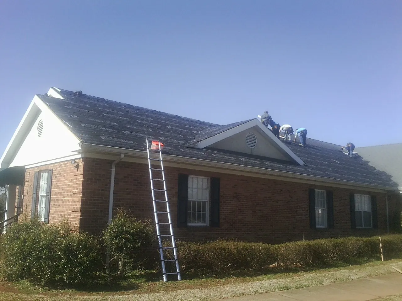 Crew tearing shingles off a brick ranch roof with a ladder set — Metal Roof Repair work by our crew in Tupper Lake
