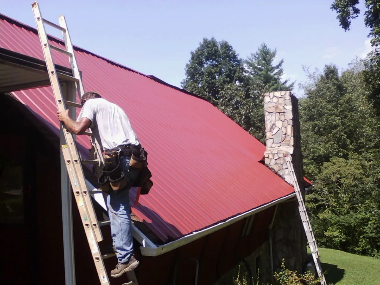 Roofer on a ladder beside a red metal roof and stone chimney — Soffit & Fascia Repair by Roofing Brothers in Tupper Lake
