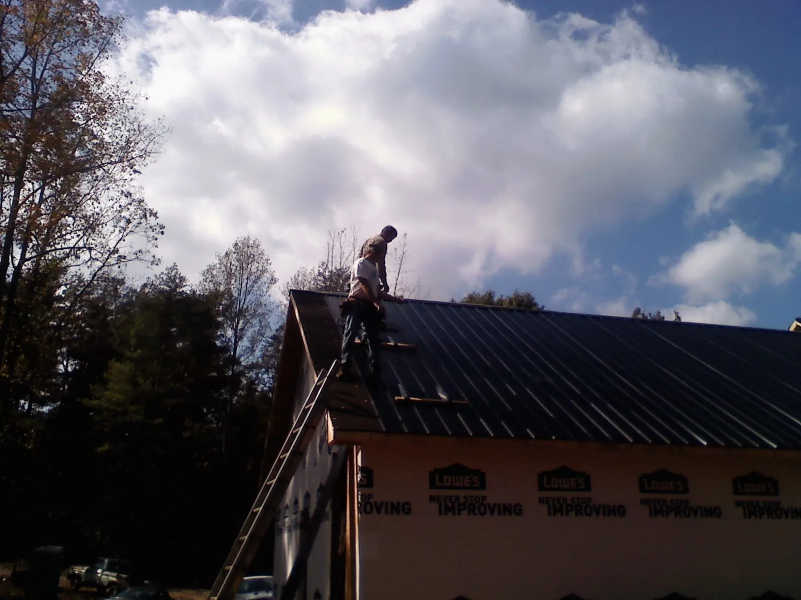 Roofer fastening dark metal panels high against a cloudy sky, from a Roof Inspection job in Tupper Lake